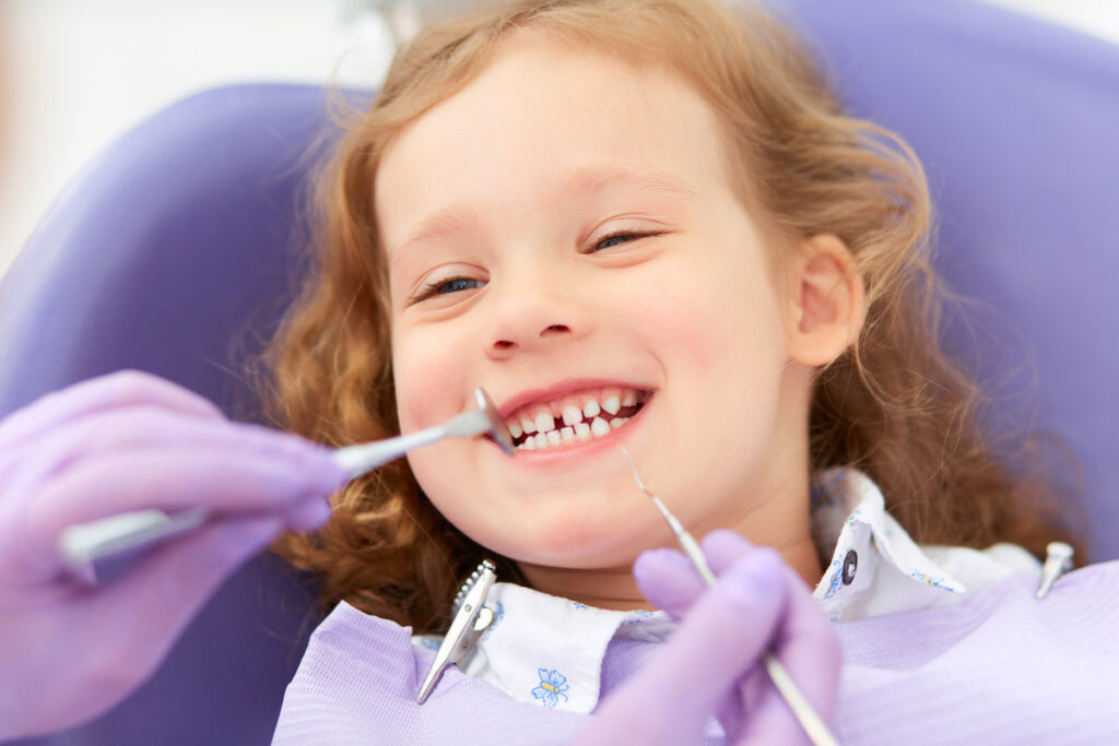 Hands of unrecognizable pediatric dentist making examination procedure for smiling cute little girl sitting on chair in hospital. Dentist office. Little girl sitting in the dentists office
