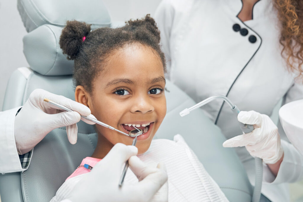 Happy afro american girl sitting in stomatologist chair with open mouth while professional dentist doing regular check up of teeth using dental probe and mirror. Female nurse assisting.