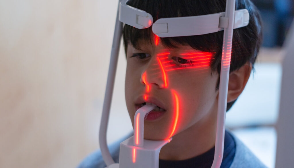 dark haired boy looking straight ahead placed on a panoramic dental x-ray machine with red lasers lighting his face with unfocused background