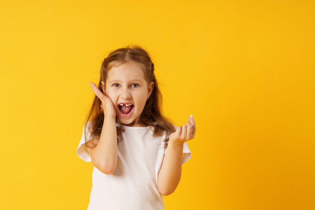 cute smiling preschool girl holds her first fallen baby tooth in her hand while standing on yellow background. growing permanent tooth is in the open mouth. . The concept of hygiene of baby teeth.
