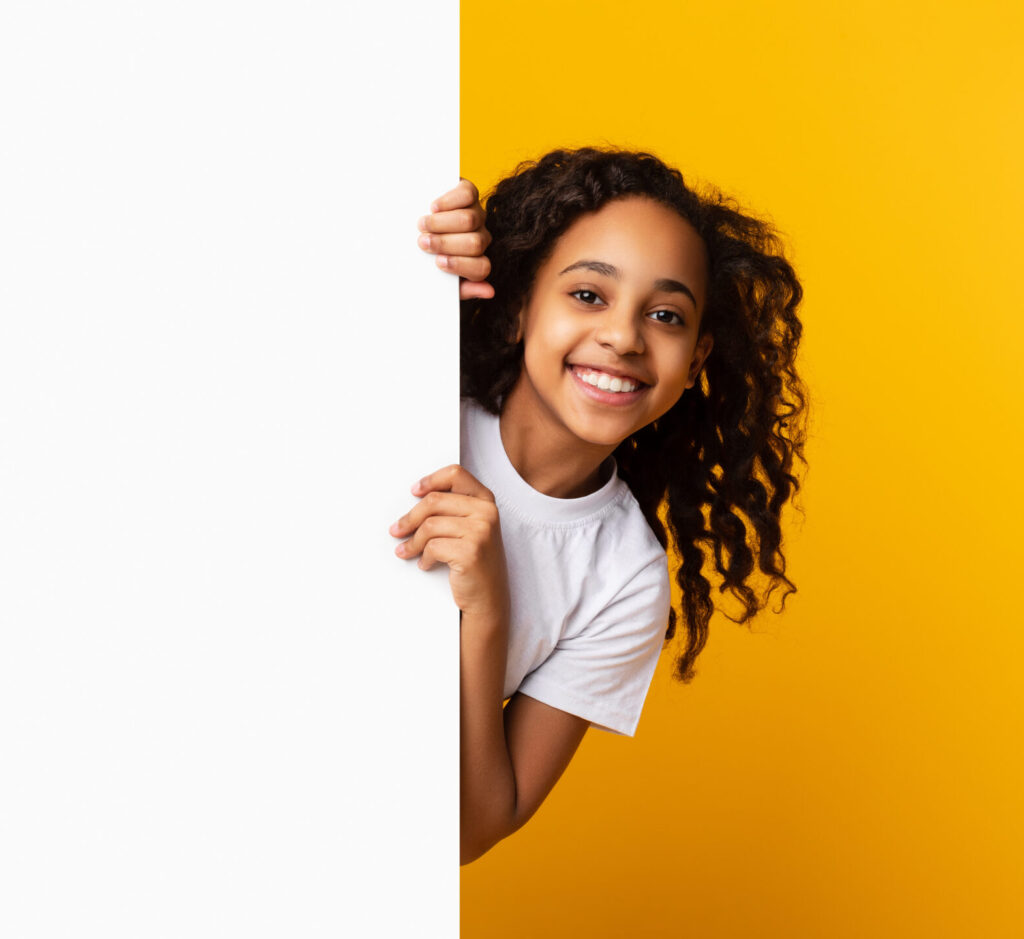 Intresting Offer. Happy Casual Black Girl Peeping Out The Side Of White Advertisement Board For Text Or Design. Smiling Cheerful Teenage Holding Billboard, Looking At Camera, Standing Over Yellow Wall