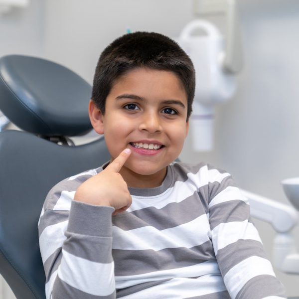 Smiling boy pointing his finger at his healthy teeth while sitting in dentist chair at clinic