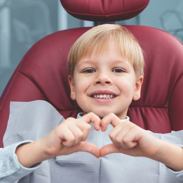 Close up portrait of a very happy boy sitting in dentist chair showing heart with his fingers and smiling at the camera, dental health and teeth care concept