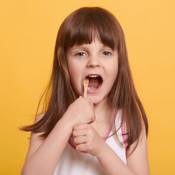 Horizontal shot of little toddler with opened mouth, holding toothbrush in both hands, brushing her first tooth, standing against yellow wall. Childhood, dental hygiene, morning procedures concept.