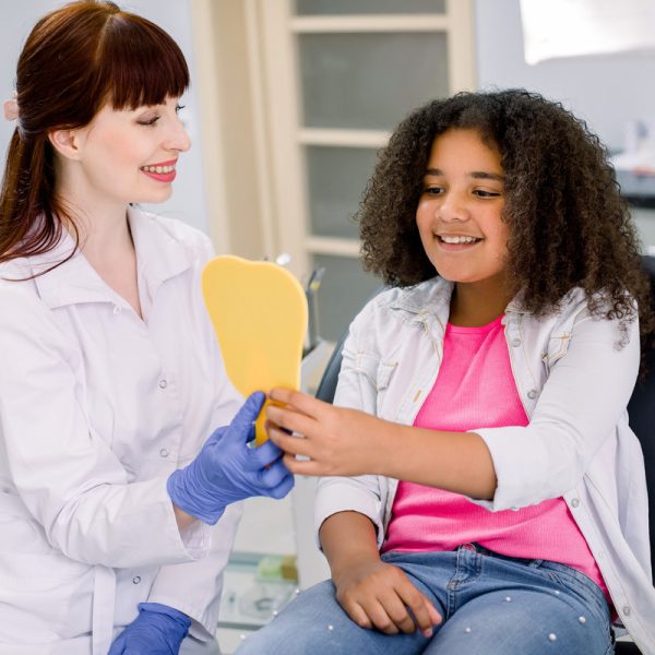 Cute happy mixed raced teen girl, looking at the mirror with toothy smile, sitting on the dental chair while smiling female dentist showing the result of treatment teeth at the dental office