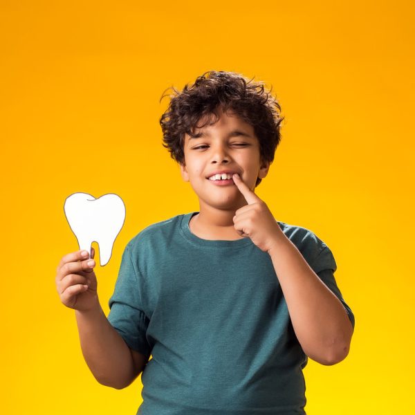 Portrait of child boy holding papercraft tooth and pointing finger at mouth, over yellow background. Dental health concept